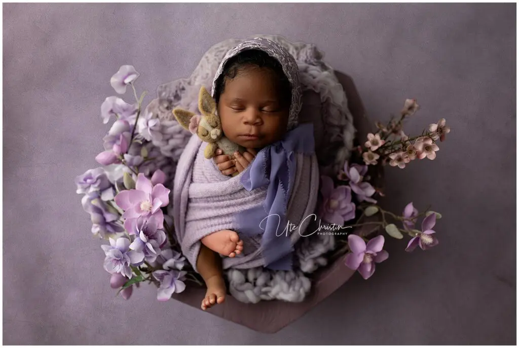 A newborn baby wrapped in a lavender blanket and bonnet sleeps peacefully, holding a small stuffed bunny. The baby is surrounded by soft purple flowers and rests on a textured purple backdrop.