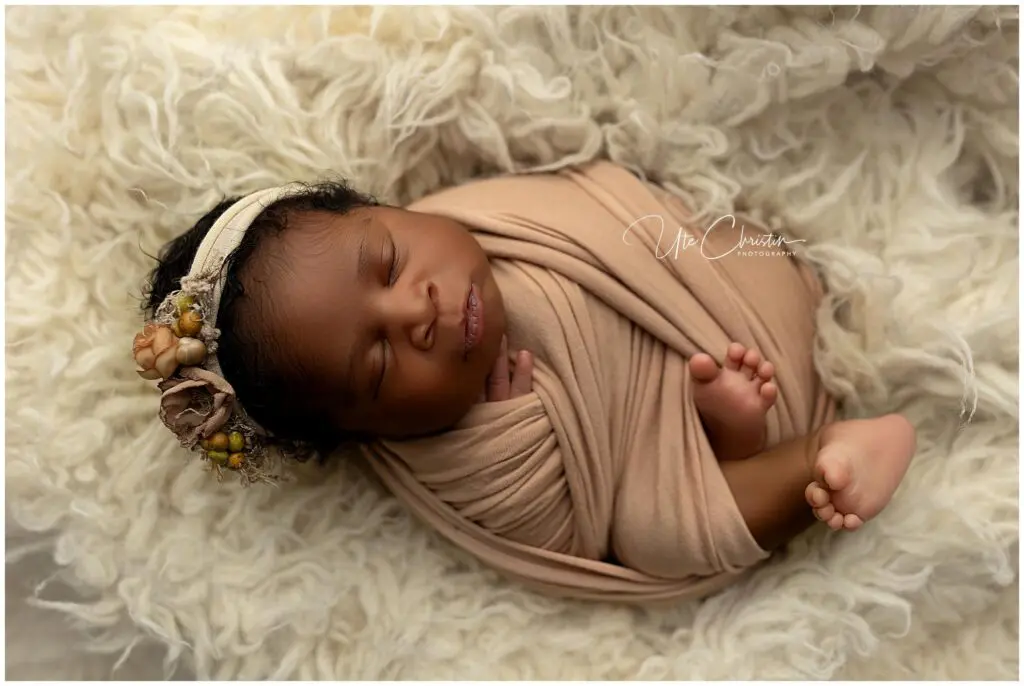 A newborn baby wrapped in a soft beige blanket sleeps peacefully on a fluffy cream-colored surface, wearing a floral headband, a serene scene perfect for sharing postpartum-insider-tips with new parents.
