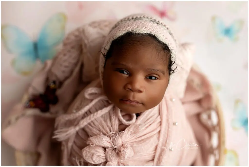 A newborn baby wrapped in a soft pink blanket and knit bonnet looks up at the camera. Perfect for capturing precious newborn photos, the background features pastel butterflies and a light, dreamy atmosphere.