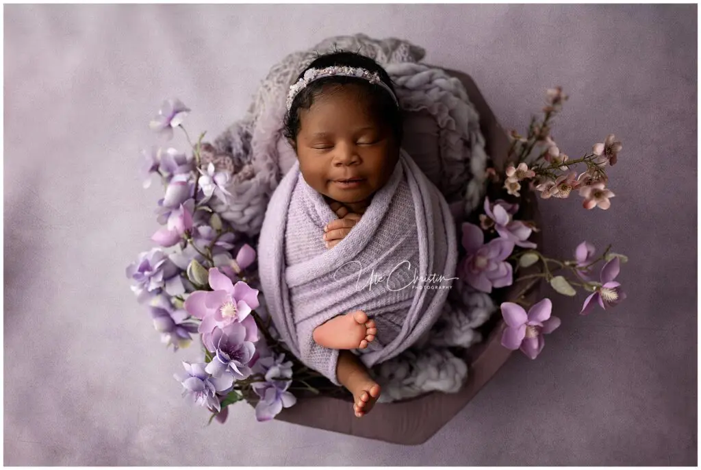 A newborn baby, wrapped in a soft lavender blanket and wearing a matching headband, sleeps peacefully in a basket surrounded by purple and pink flowers on a lavender background.