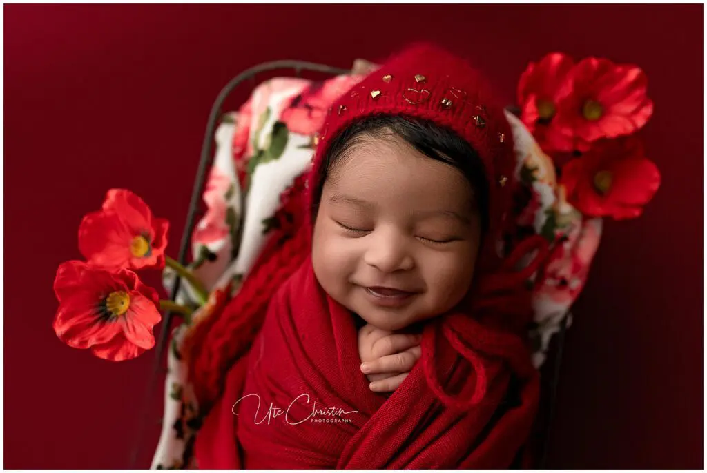A smiling newborn wrapped in a red blanket and hat, lying on a floral cushion with red flowers around, against a deep red background.