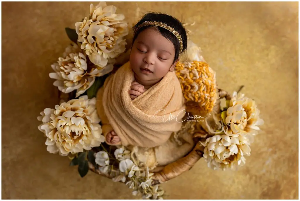A newborn baby wrapped in a soft beige blanket sleeps peacefully in a round basket, surrounded by large cream-colored flowers on a golden background. Perfect for newborn photos CT, the baby wears a delicate headband.