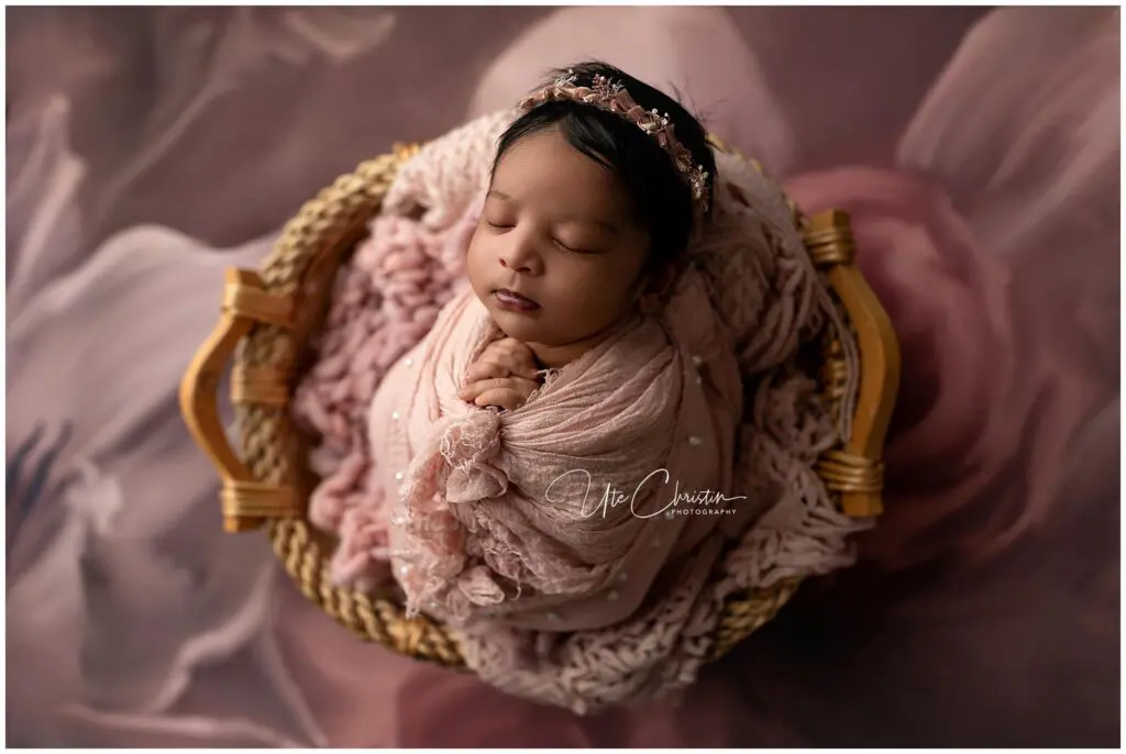 A newborn baby wrapped in a soft pink blanket sleeps peacefully in a woven basket, wearing a pink floral headband, with a blurred floral background.