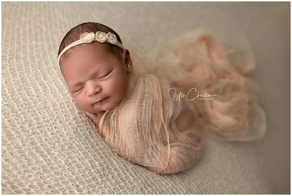 A newborn baby sleeps peacefully, swaddled in a soft beige wrap with a delicate flower headband, lying on a textured cream blanket. The background is soft and neutral, creating a gentle, serene atmosphere.