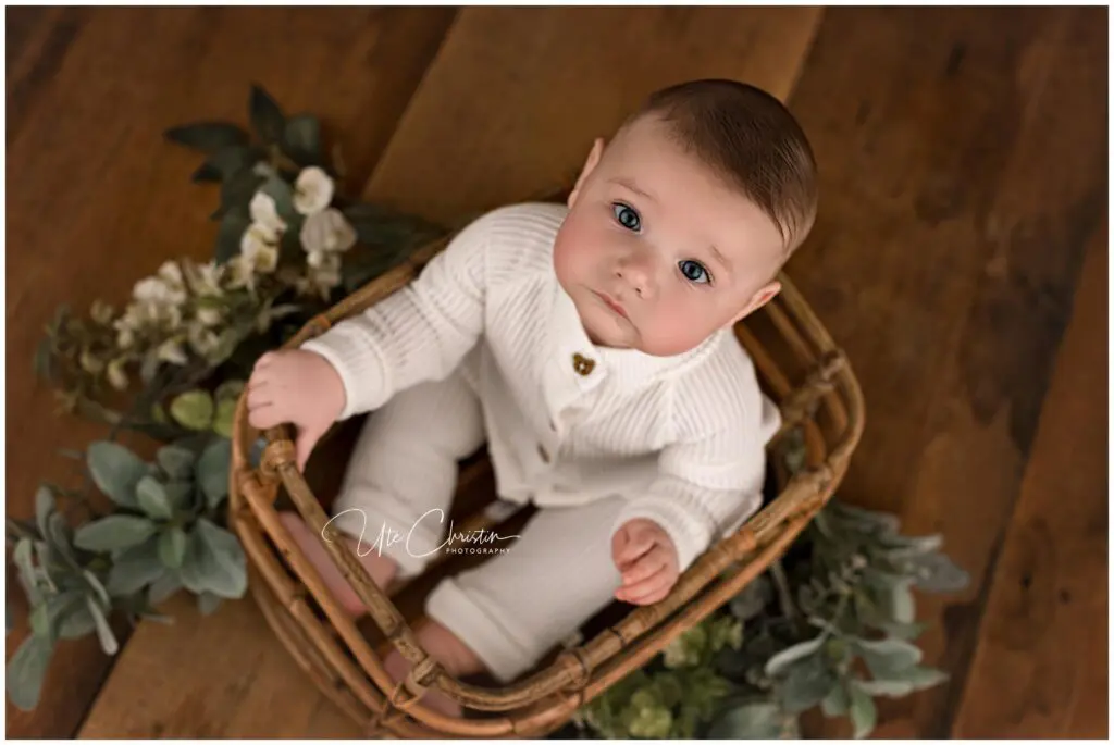 A baby with blue eyes, wearing a white knit outfit, sits in a wicker basket surrounded by green foliage and flowers on a wooden floor, looking up at the camera.