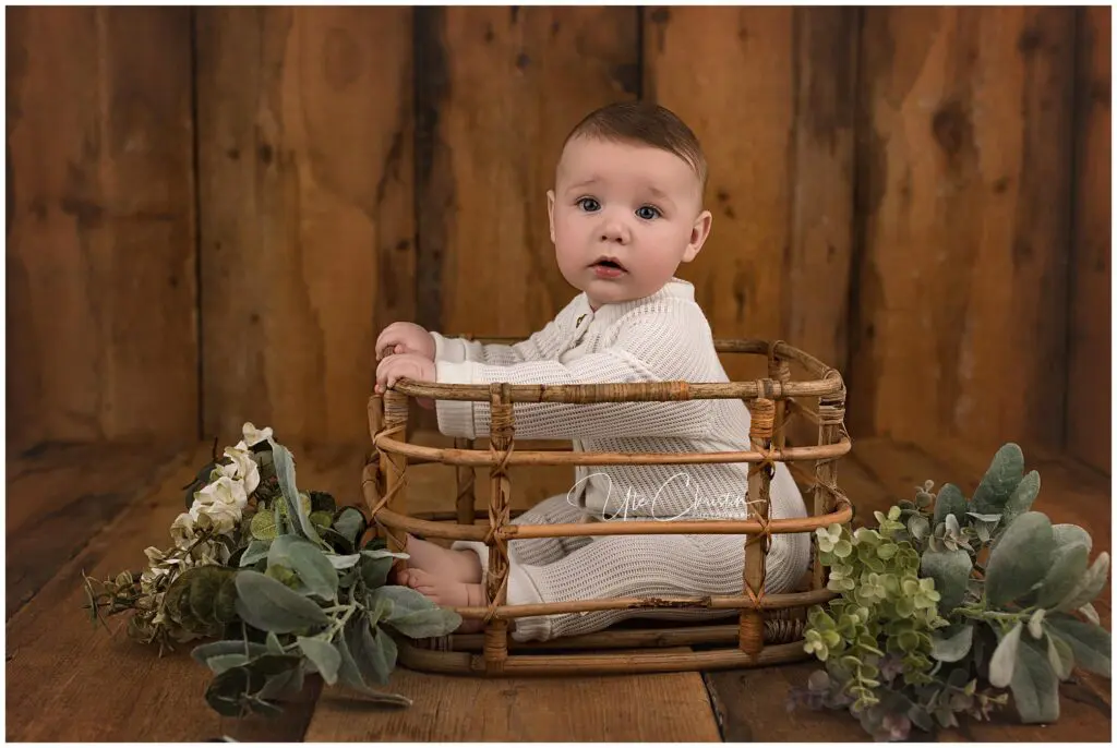 A baby wearing a cream-colored outfit sits inside a woven basket, surrounded by green and white artificial flowers, against a wooden background. The baby looks toward the camera with a curious expression.