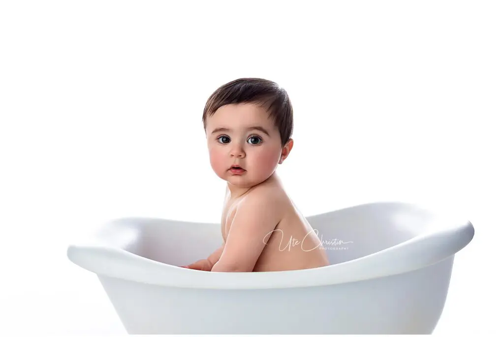 A baby with dark hair and big eyes sits in a white tub, looking over their shoulder towards the camera against a plain white background.