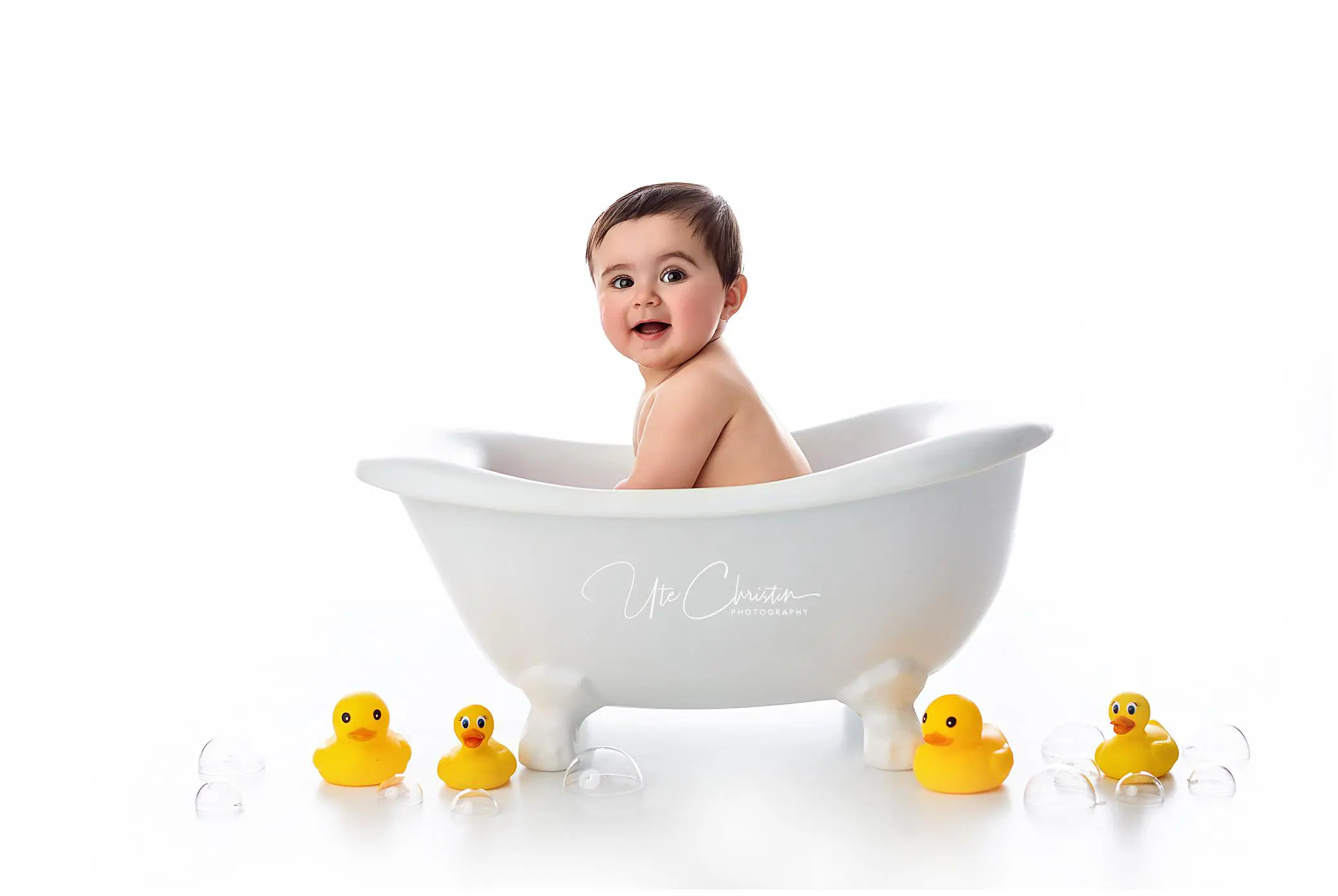A smiling baby sits in a small white bathtub surrounded by several yellow rubber ducks and clear plastic bubbles, against a white background.