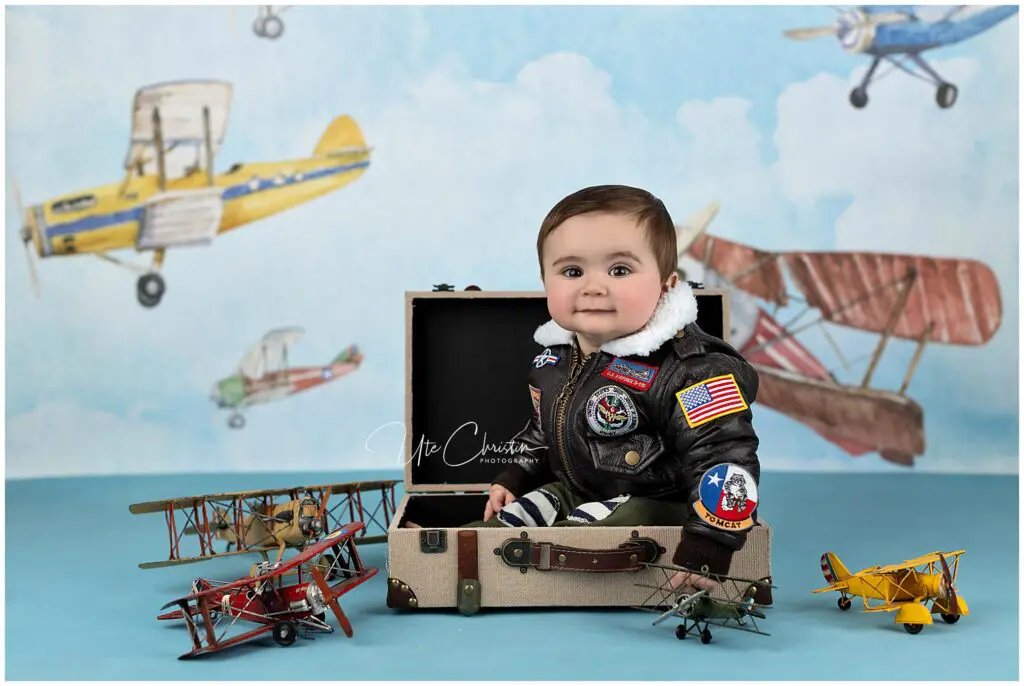 A smiling baby in a leather aviator jacket sits in an open suitcase, surrounded by toy biplanes with a sky and airplanes painted on the backdrop.