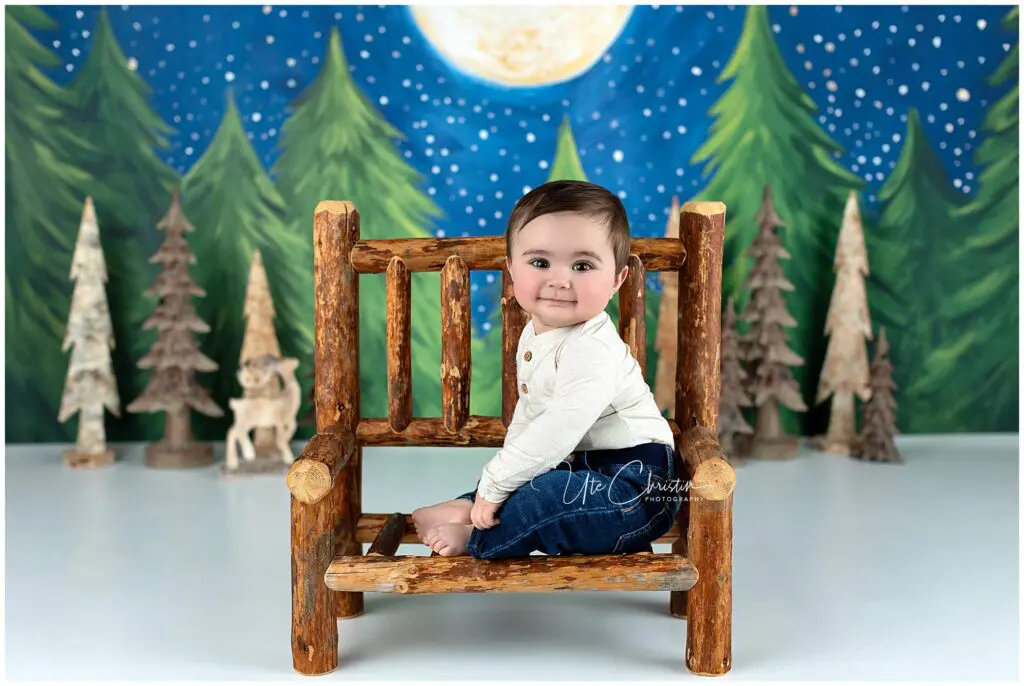 A smiling baby with dark hair sits sideways on a rustic wooden bench, wearing a cream shirt and jeans. Behind, there’s a painted backdrop of a night sky, full moon, pine trees, and painted animal figures.