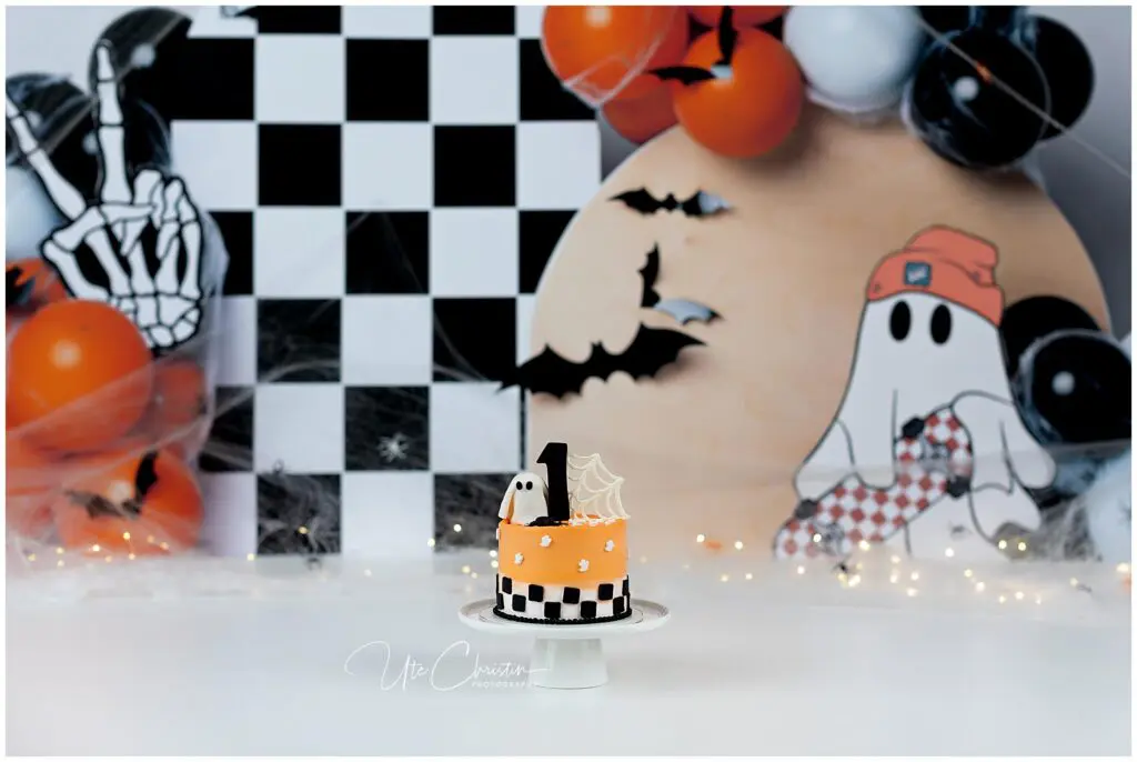 A small orange Halloween-themed cake with a black number 1 topper and ghost decoration sits on a stand. The background features balloons, spider webs, bats, and a cartoon ghost illustration.