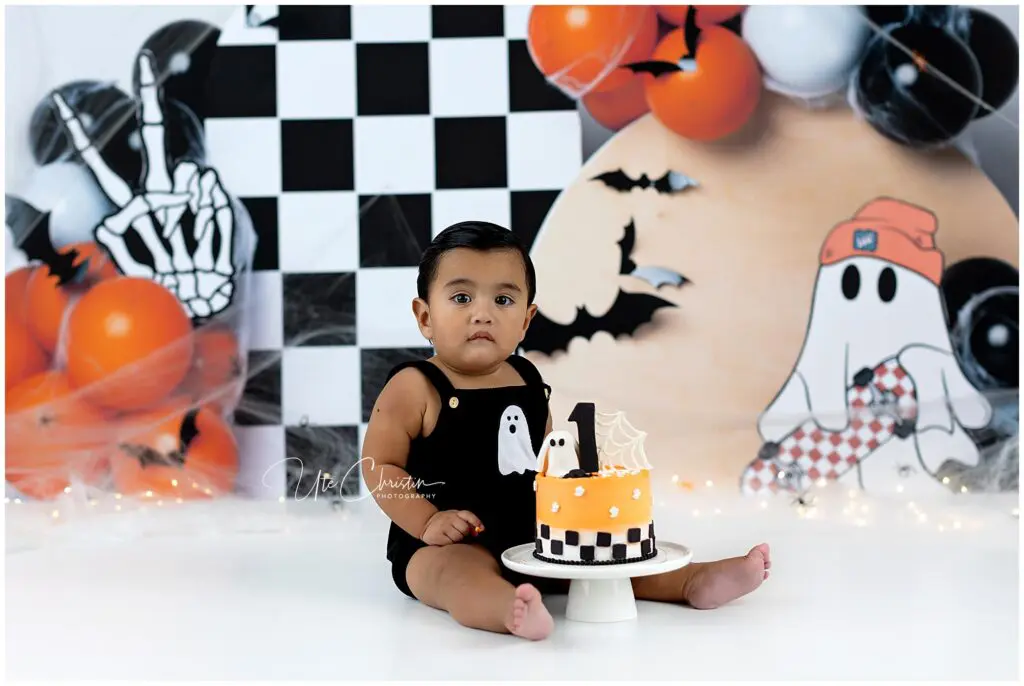 A toddler in a black ghost romper sits on the floor with a Halloween-themed cake. The background features black, white, and orange balloons, bats, and a ghost illustration.