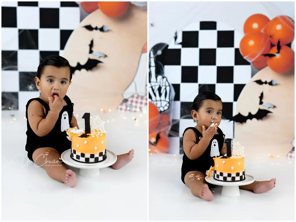 A young child in a black outfit sits on the floor eating an orange Halloween-themed cake with a 1 topper. The backdrop has black and white checks, bats, and orange balloons.