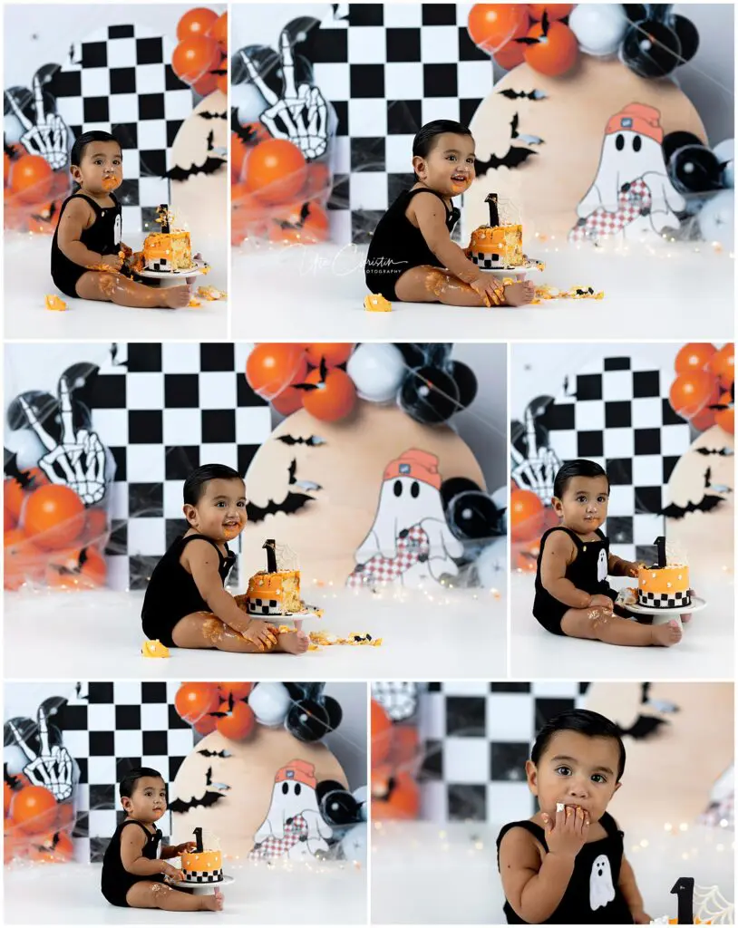 A toddler in a black outfit has a Halloween-themed cake smash. The background features balloons, bats, and cartoon ghosts with a checkered pattern. The child is sitting and playing with the cake, smiling and looking curious.