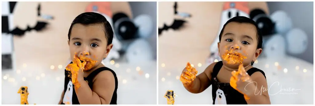 A baby with dark hair wearing a black outfit messily eats orange cake, getting icing on their face and hands. Halloween decorations are blurred in the background.