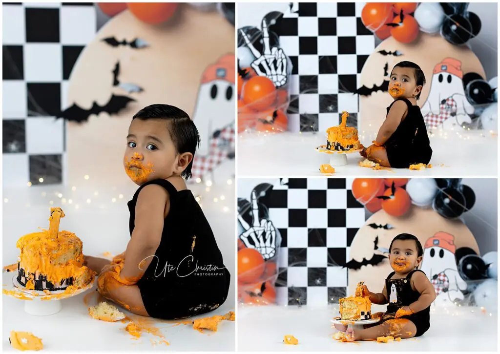 A toddler in a black outfit sits on the floor with an orange frosted cake, surrounded by Halloween-themed decorations. The child has cake on their face and hands, smiling in a playful, messy cake smash photo session.