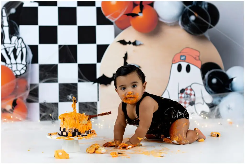 A baby in a black outfit crawls on the floor with orange cake smeared on their face and body. The background has Halloween decorations, including balloons, bats, and a cartoon ghost.
