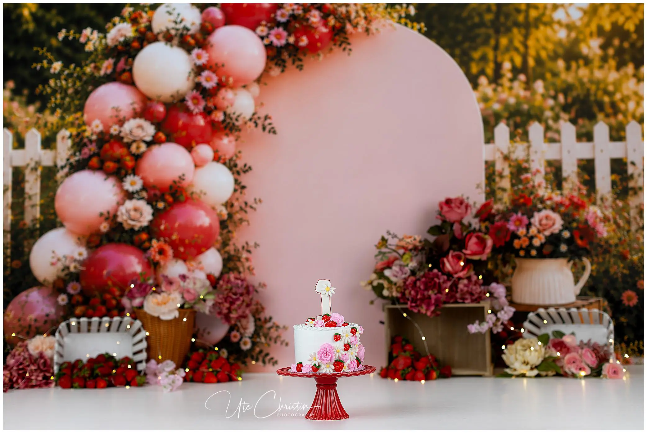 A small white cake with pink flowers sits on a red stand, surrounded by baskets of strawberries, pink balloons, and flower arrangements with a white picket fence and greenery in the background.