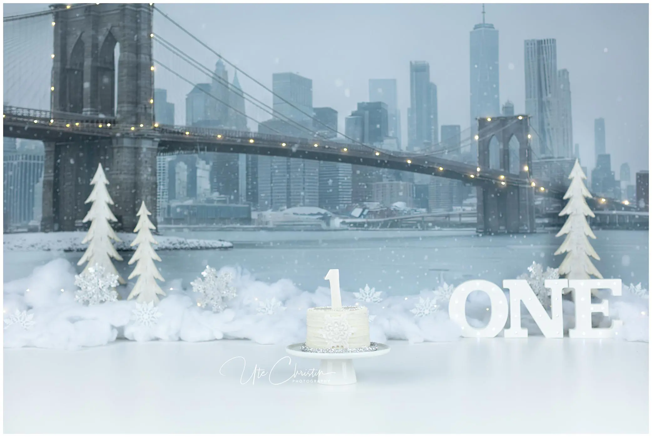 A small white cake with a number 1 topper sits on a table. Snowy decorations, white trees, and the word “ONE” surround it. In the background is the Brooklyn Bridge and a city skyline in winter.