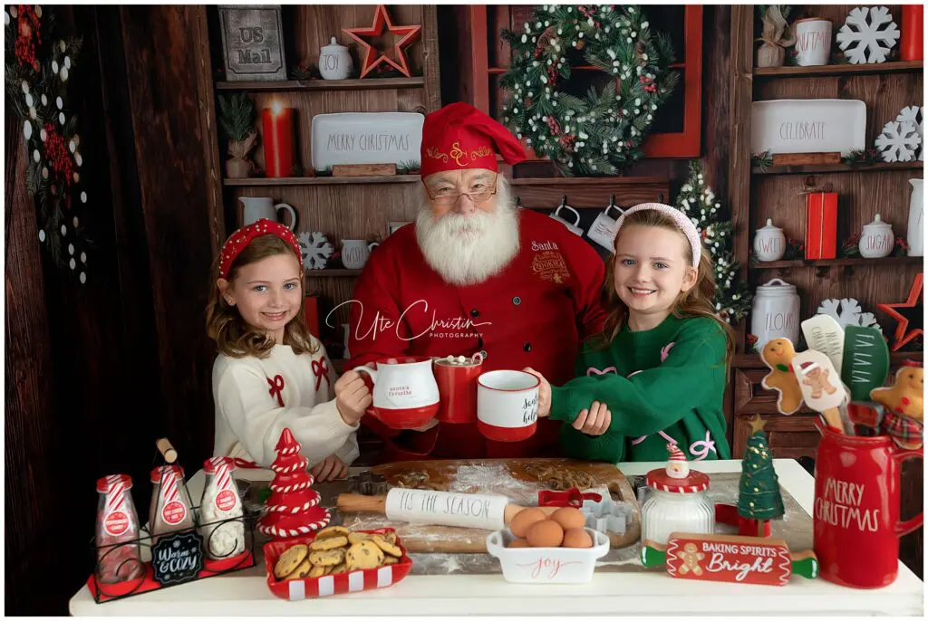 Two smiling children and a man dressed as Santa Claus hold mugs in a festive Christmas kitchen, surrounded by cookies, eggs, hot cocoa, and holiday decor. Shelves display jars, wreaths, and holiday signs in the background.