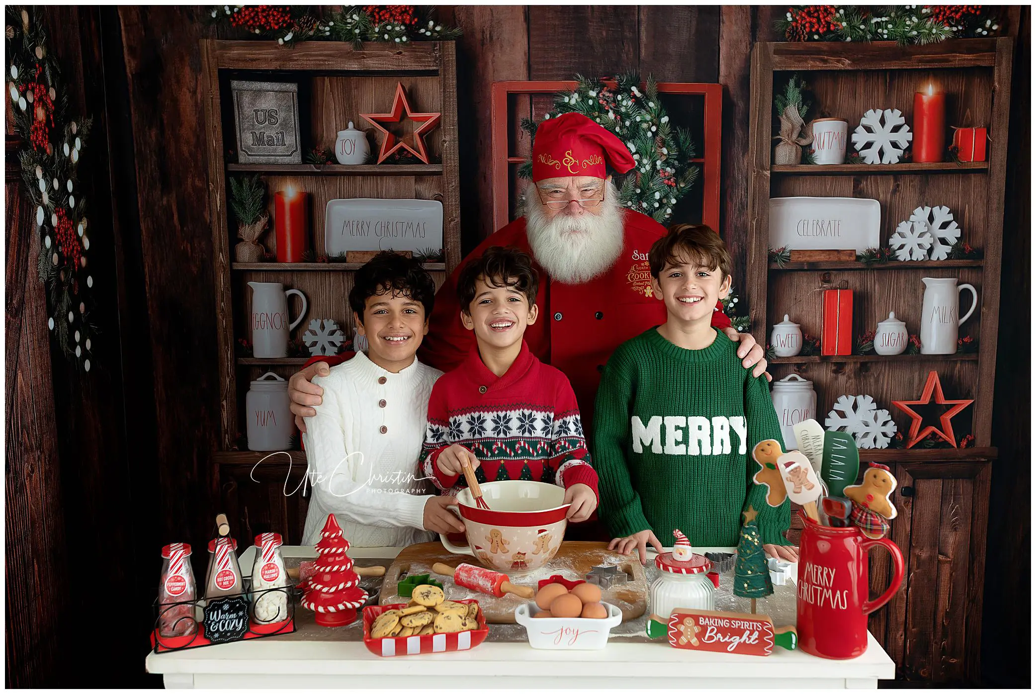 Three smiling boys stand in front of a festive Christmas kitchen setup with Santa Claus, surrounded by holiday decorations, cookies, milk bottles, and baking supplies on a white table.