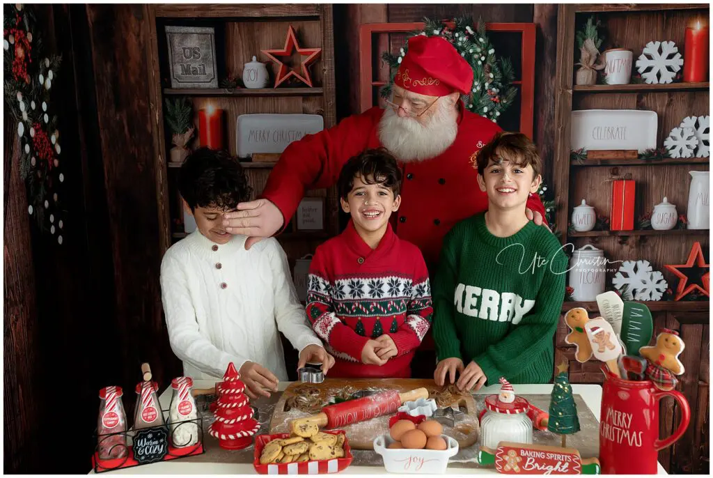 Santa stands behind three smiling children in holiday sweaters at a festive kitchen counter with cookies, eggs, and Christmas decorations. The children look happy as they prepare holiday treats together.
