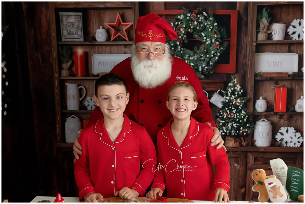 Santa Claus stands behind two smiling children in matching red pajamas. They are in a festive kitchen decorated with wreaths, wooden shelves, and holiday treats, creating a warm Christmas atmosphere.