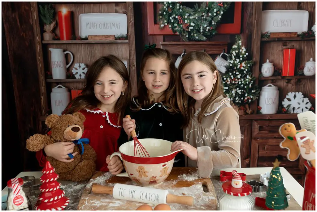 Three young girls in festive pajamas smile in a decorated kitchen, mixing batter in a gingerbread bowl. One holds a teddy bear, and Christmas decorations, cookies, and a sign that reads Tis the Season are on the table.