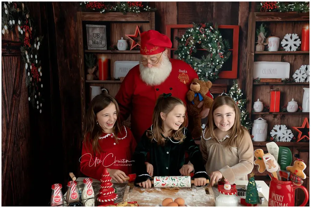 Santa stands behind three smiling children at a festive kitchen table, helping them bake Christmas treats. The background is decorated with wreaths, jars, holiday signs, and festive decor.