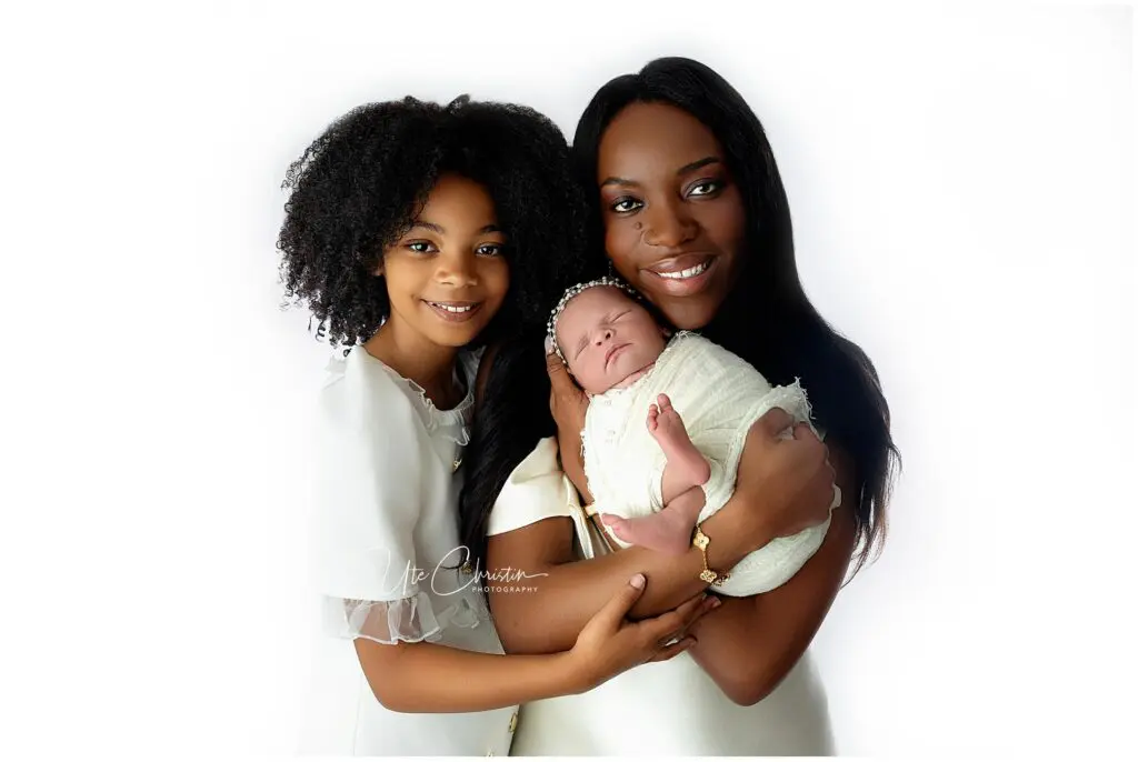 A woman holds a swaddled newborn while standing next to an older girl. All three are smiling, dressed in white, and posed against a bright white background.