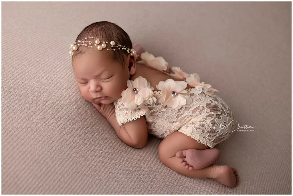 Newborn baby sleeping on a textured beige blanket, wearing a lace romper with flower accents and a pearl headband, posed on their side with hands under their cheek.