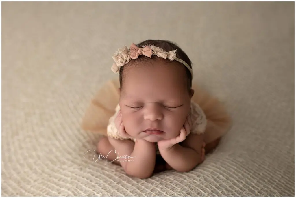 Newborn baby with a floral headband sleeps with chin resting on hands, lying on a soft beige blanket and wearing a peach tulle outfit.