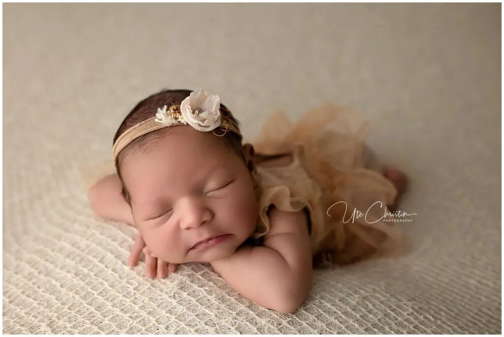 Newborn baby sleeping on stomach on a cream textured blanket, wearing a beige outfit and a headband with a flower.
