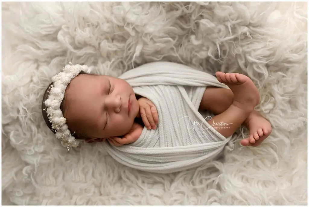 Newborn baby sleeping peacefully, swaddled in a white blanket with a delicate floral headband, lying on a soft, fluffy white surface.
