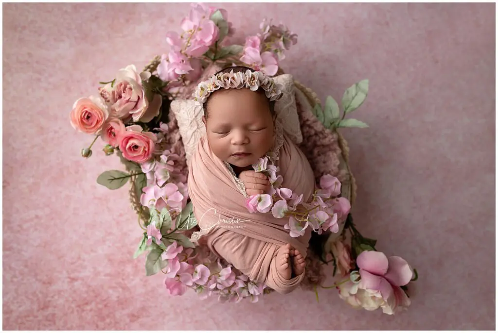 Newborn baby wrapped in a soft pink blanket, sleeping peacefully in a basket surrounded by pink flowers, with a floral headband, on a pink textured background.