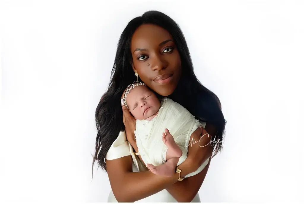 A woman with long dark hair cradles a sleeping newborn wrapped in a white blanket, both facing the camera against a white background.