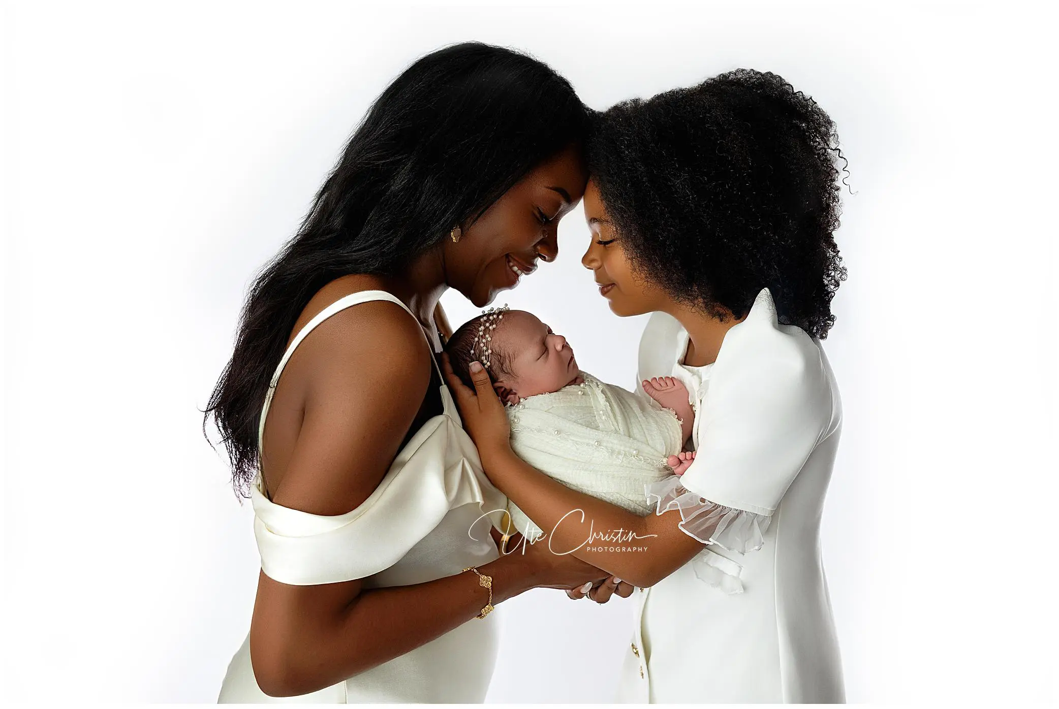 Two women in white gently touch foreheads while smiling at a swaddled newborn cradled between them, against a white background.