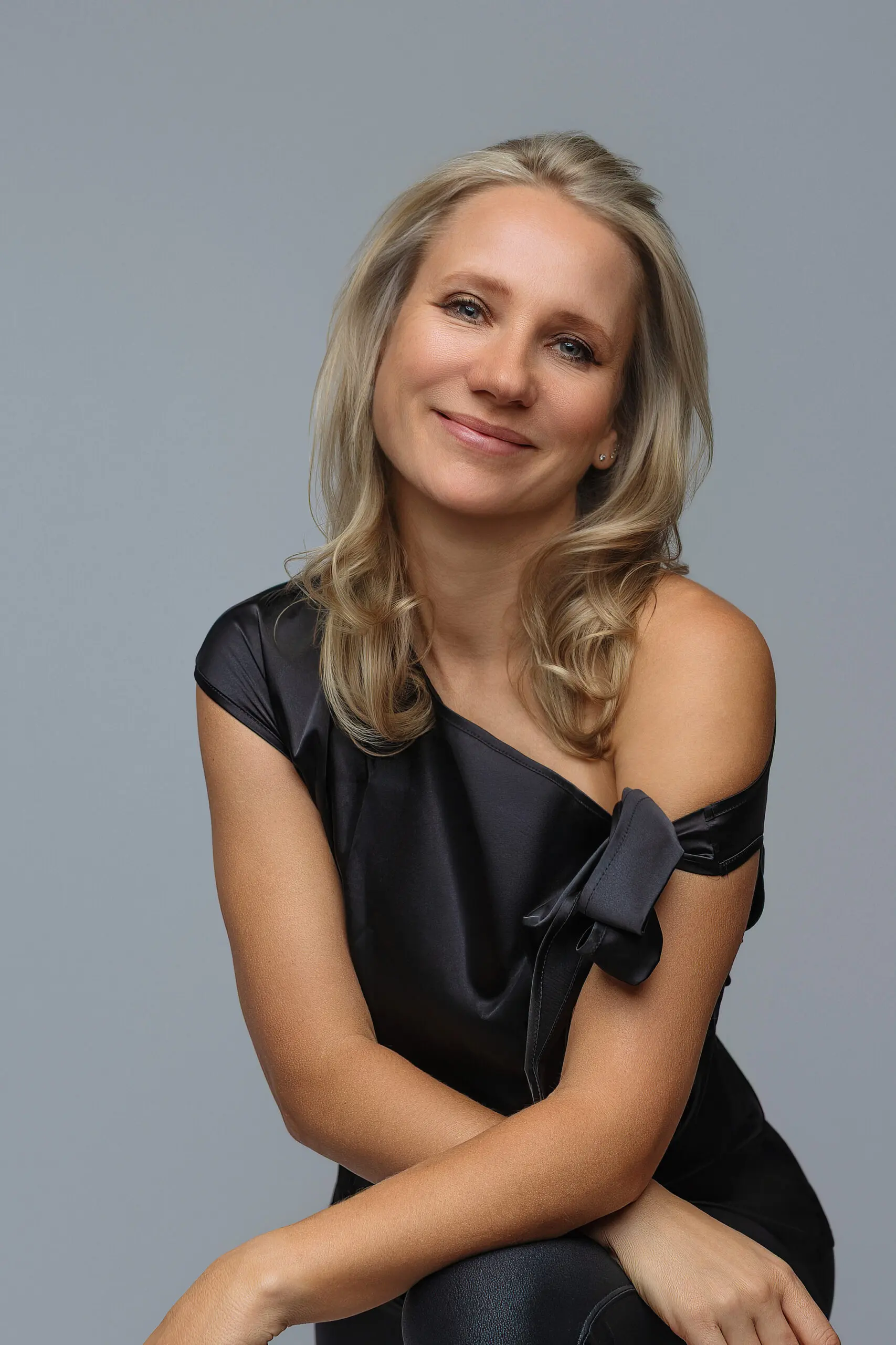 A Milford photographer with blonde hair and light skin smiles gently, wearing a black, off-the-shoulder dress against a plain gray background.