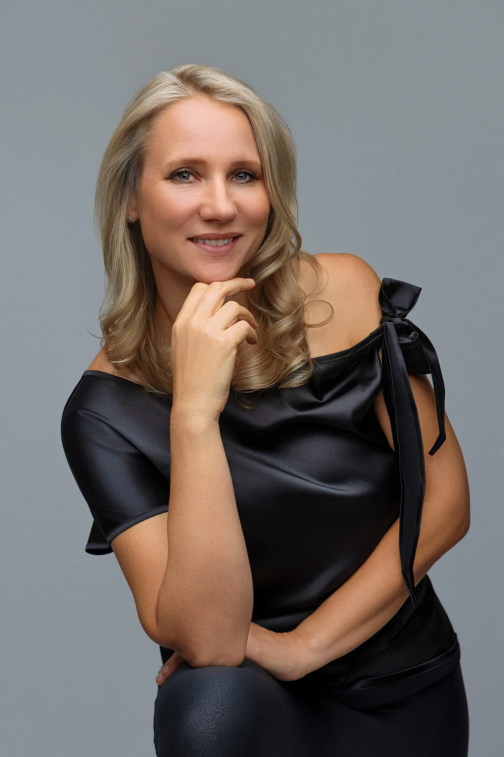 Smiling woman with long blonde hair in a black off-shoulder top, resting her chin on her hand, posing against a plain gray background.