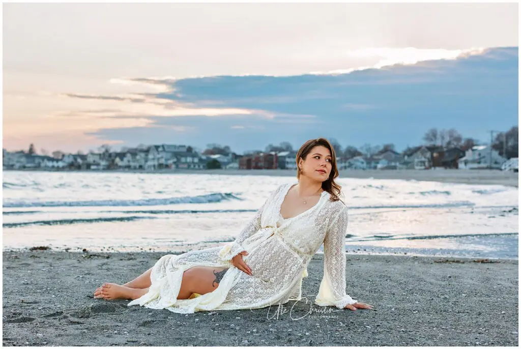 A pregnant woman in a white lace dress sits on the beach at sunset, gazing thoughtfully to the side with one hand resting on her belly. Houses and gentle waves are visible in the background.