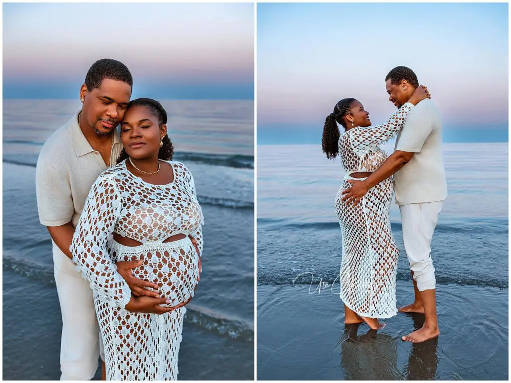 A couple stands on a beach at sunset. The woman, visibly pregnant, wears a white crochet dress. The man, in light clothing, embraces her affectionately as they stand barefoot in shallow water.