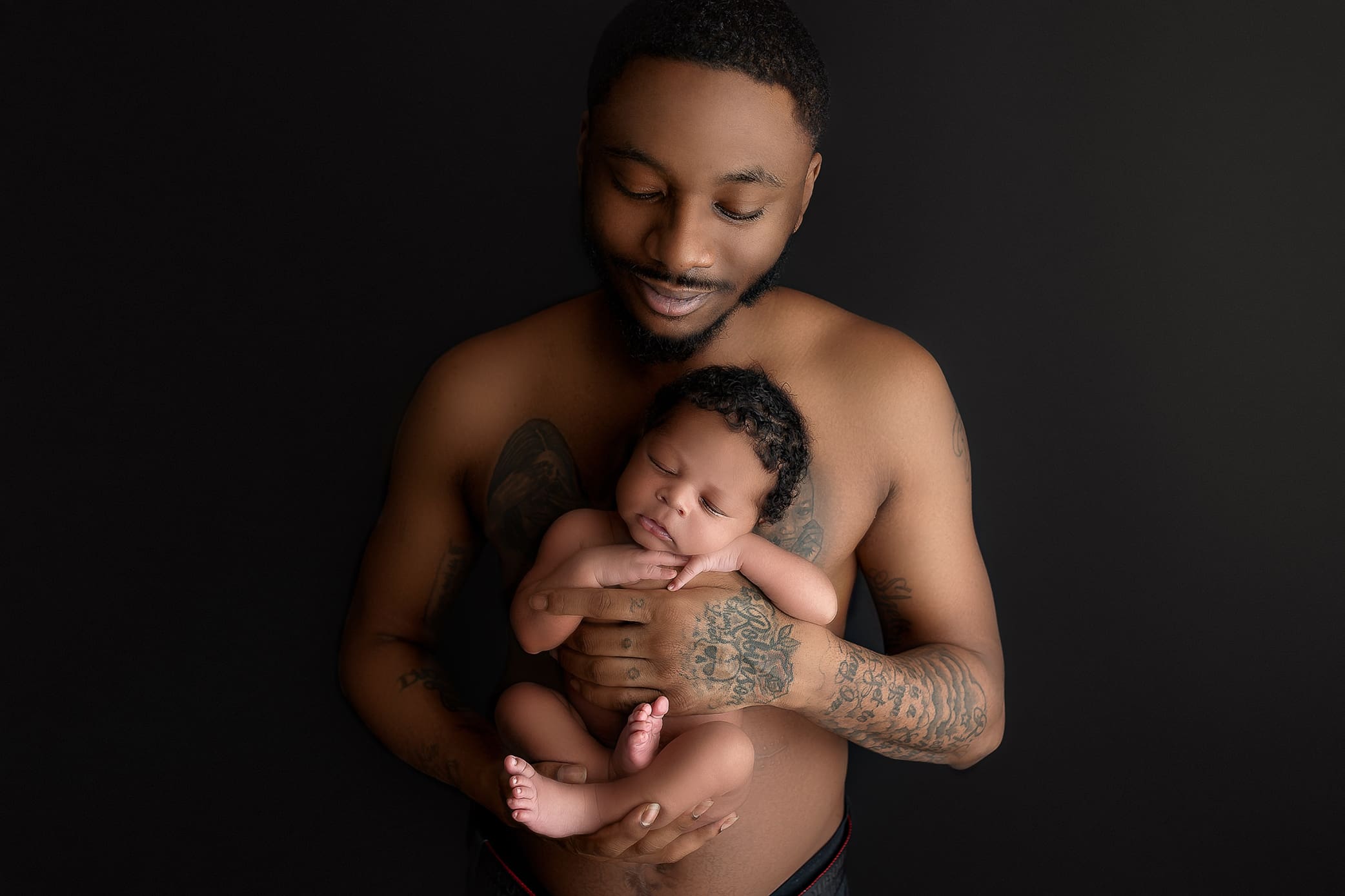 A shirtless, tattooed man gently cradles a sleeping baby in his arms, set against a dark background. Captured by a top Connecticut newborn photographer, the man looks down at the baby with a serene expression.