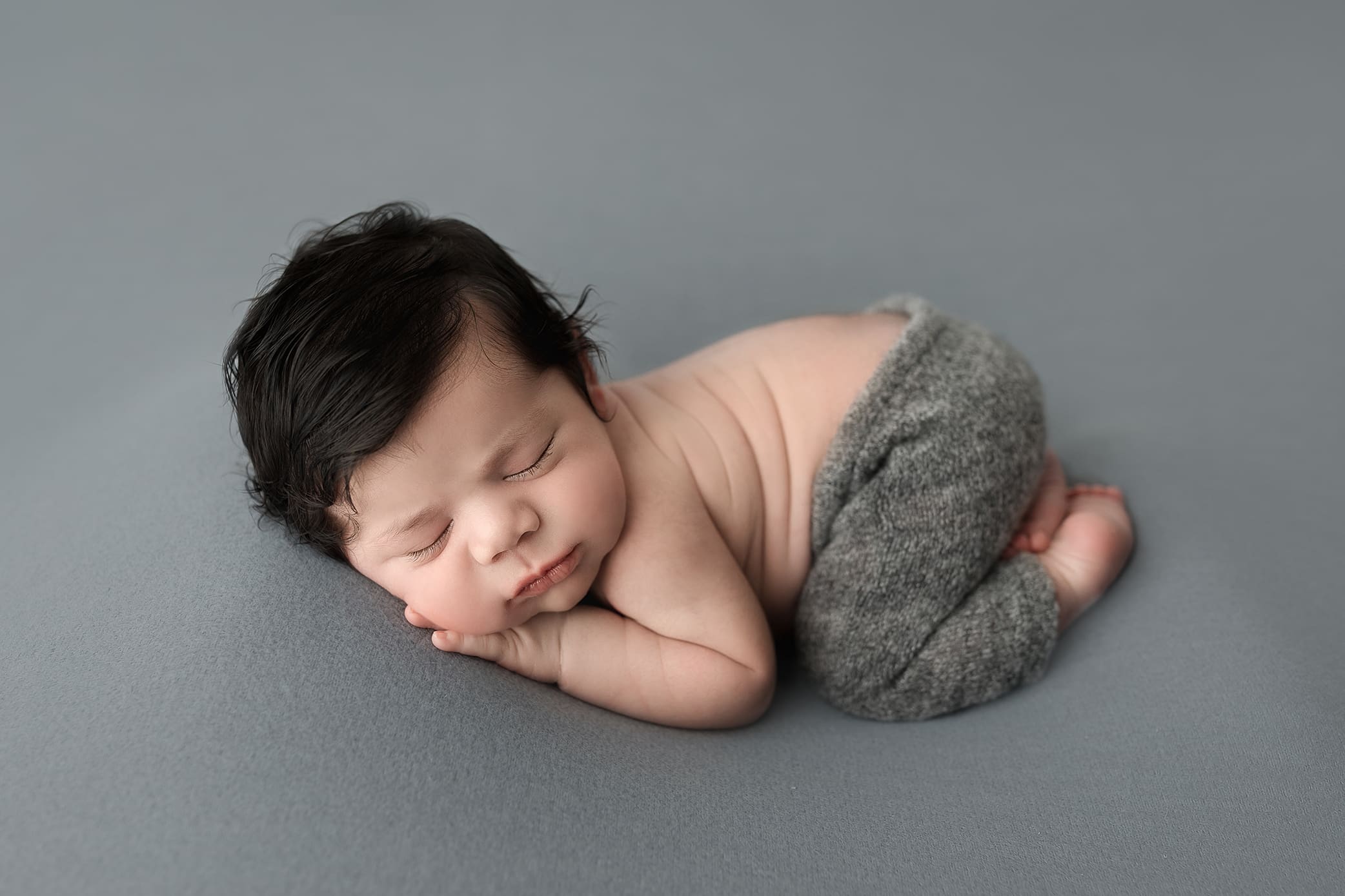 A sleeping baby with dark hair lies curled up on a gray blanket. The baby is wearing soft gray pants and has their head resting on their hands, looking peaceful and content.