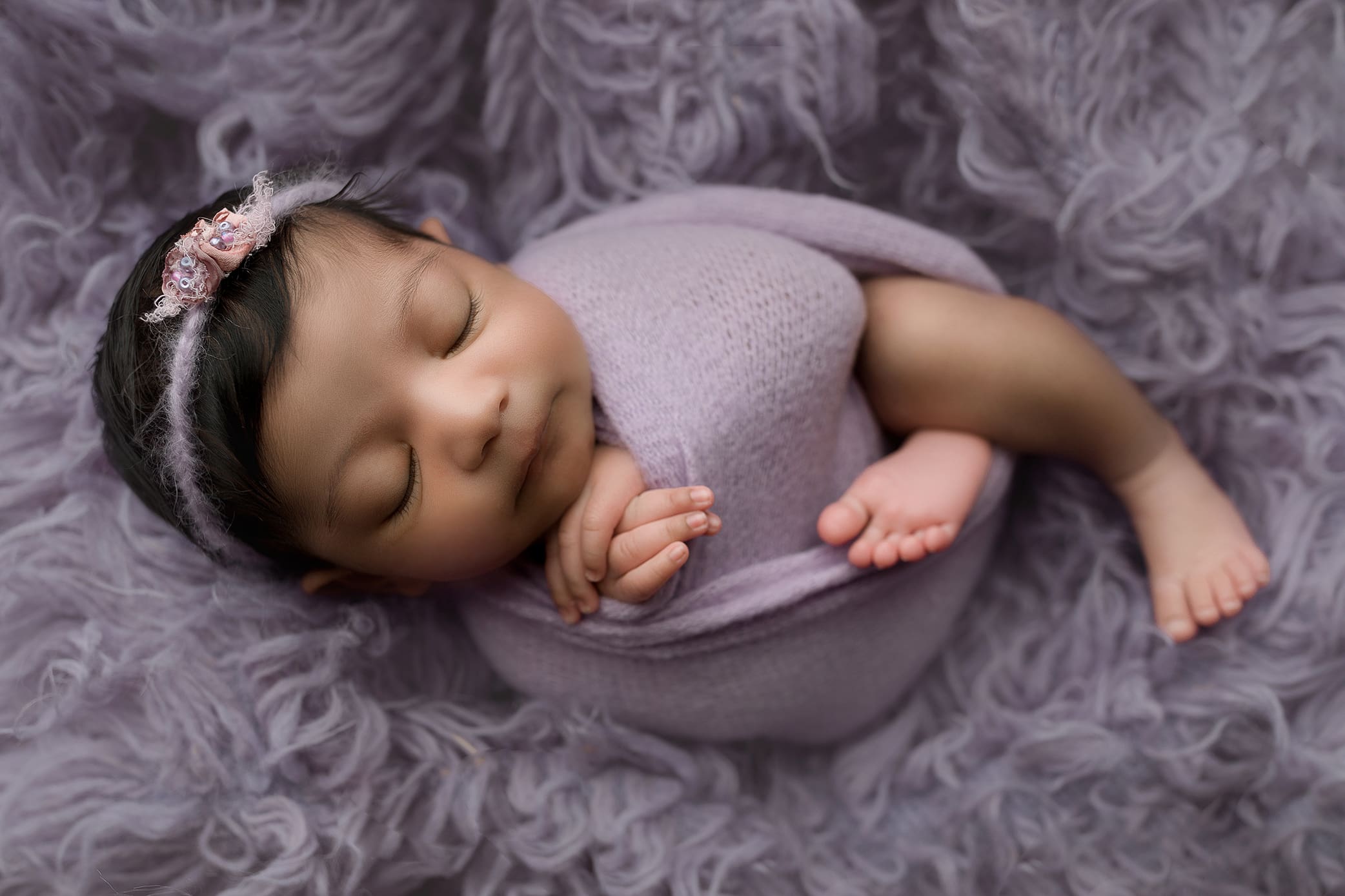 A sleeping baby, captured by a skilled baby photographer, is swaddled in a soft lavender blanket with a matching headband lying peacefully on a fluffy purple surface. The babys dark hair and tiny feet are visible, creating a serene and cozy scene.