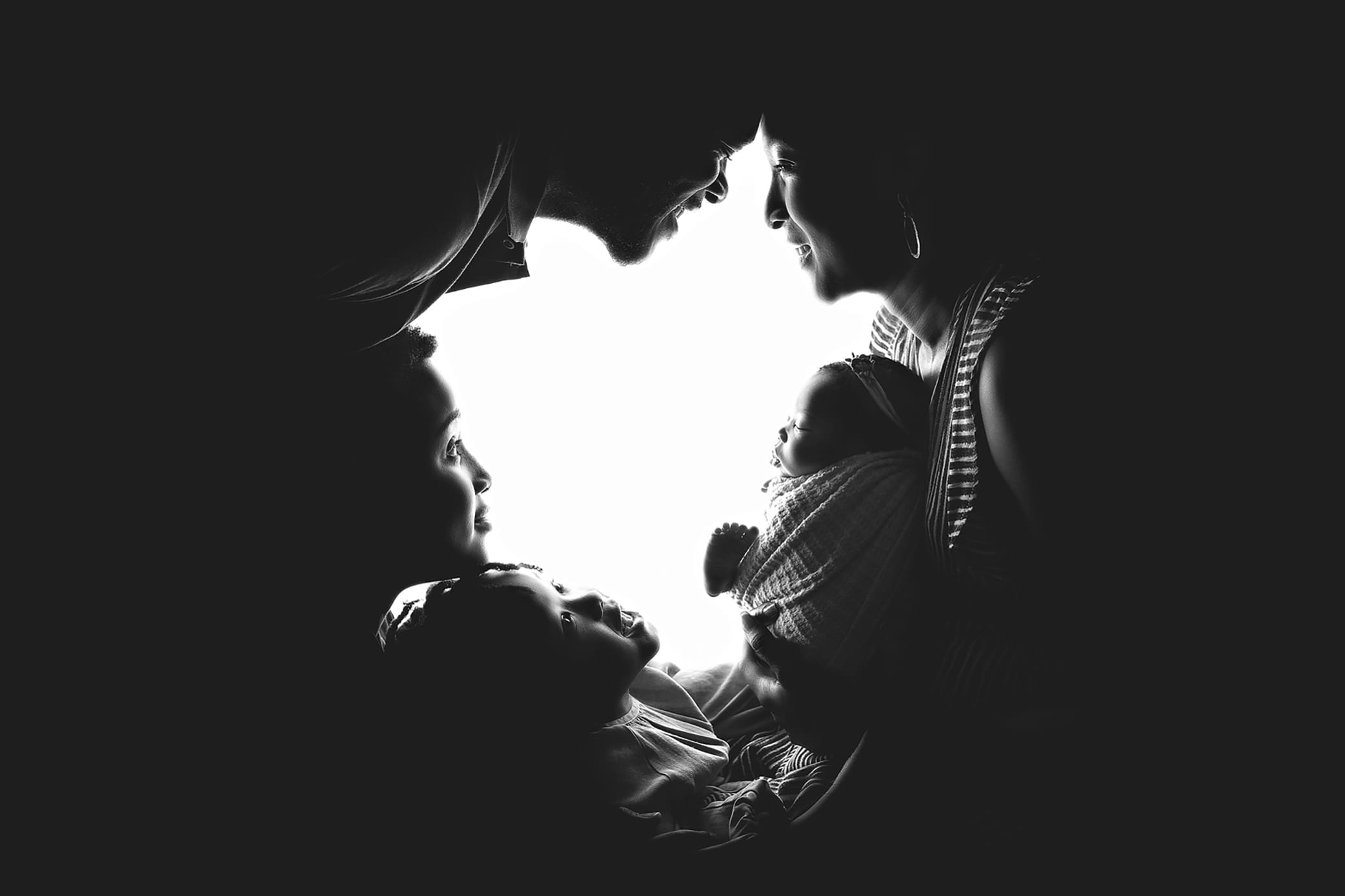 Silhouette of a family; a couple leans together touching foreheads while holding a baby. Two children stand below them, looking up, creating a heart-like shape with their faces and bodies against a bright background.