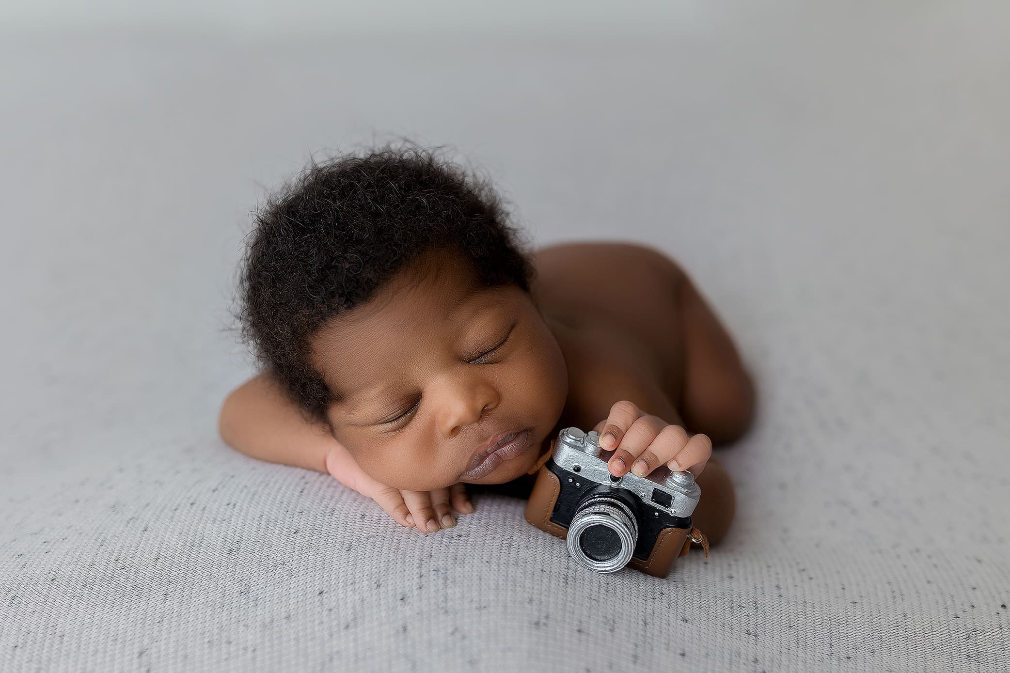 A sleeping baby is lying on their stomach, resting their head on their hands, clutching a small toy camera. Captured by a top Connecticut newborn photographer, the serene moment unfolds against a soft, neutral gray backdrop.