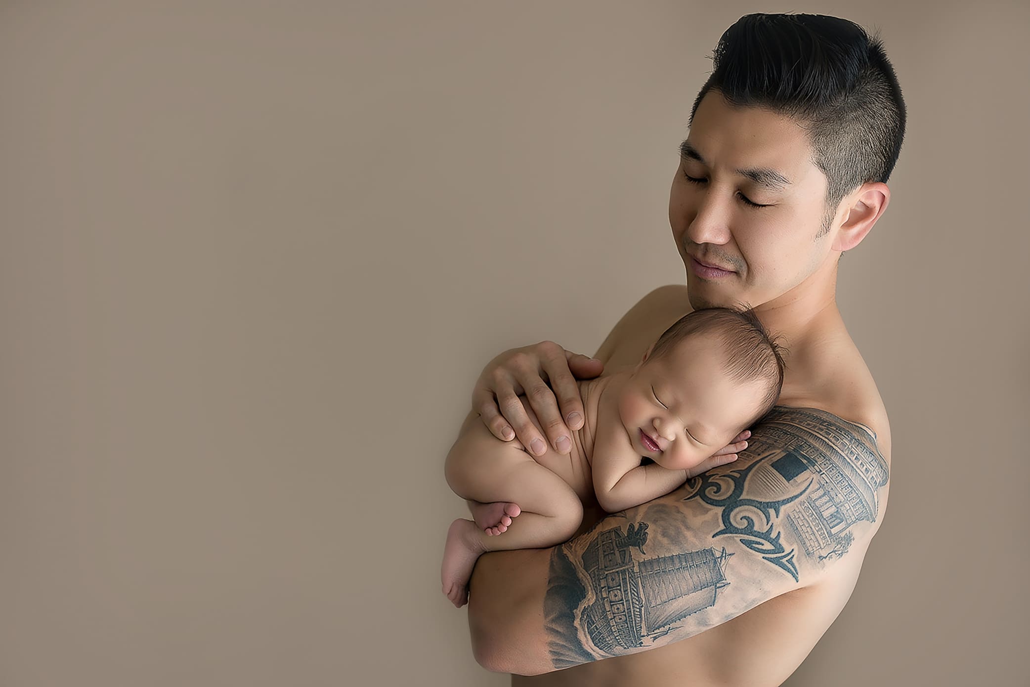 A man with tattoos on his arm, captured by a Connecticut newborn photographer, lovingly holds a sleeping baby against his chest. Both are shirtless against a plain, soft beige background. The mans eyes are closed, exuding peace and contentment.