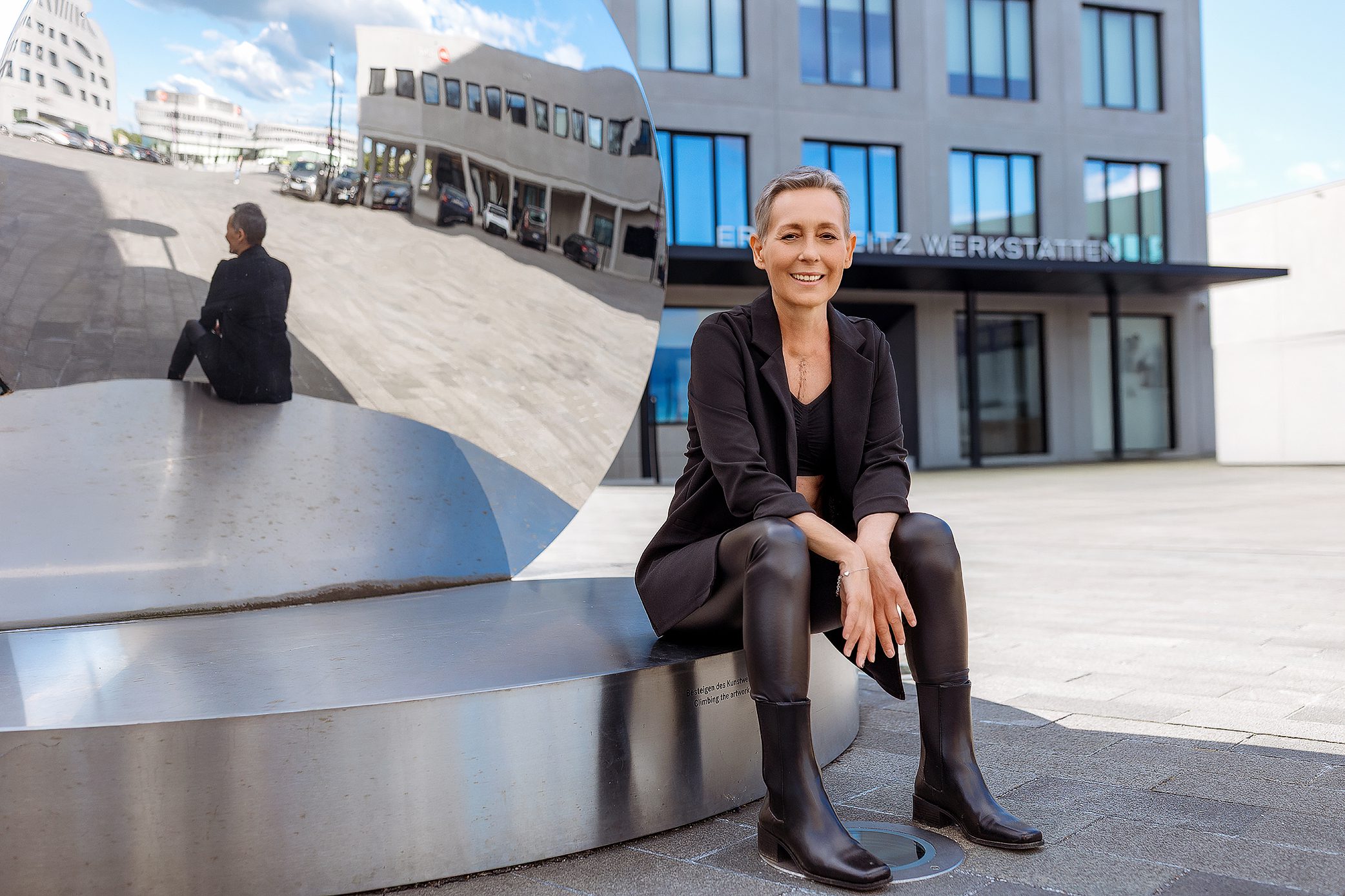 A woman with short hair, dressed in black, sits on a shiny metal sculpture outdoors. Her reflection is visible in the curved surface. Modern buildings and blue sky are in the background.