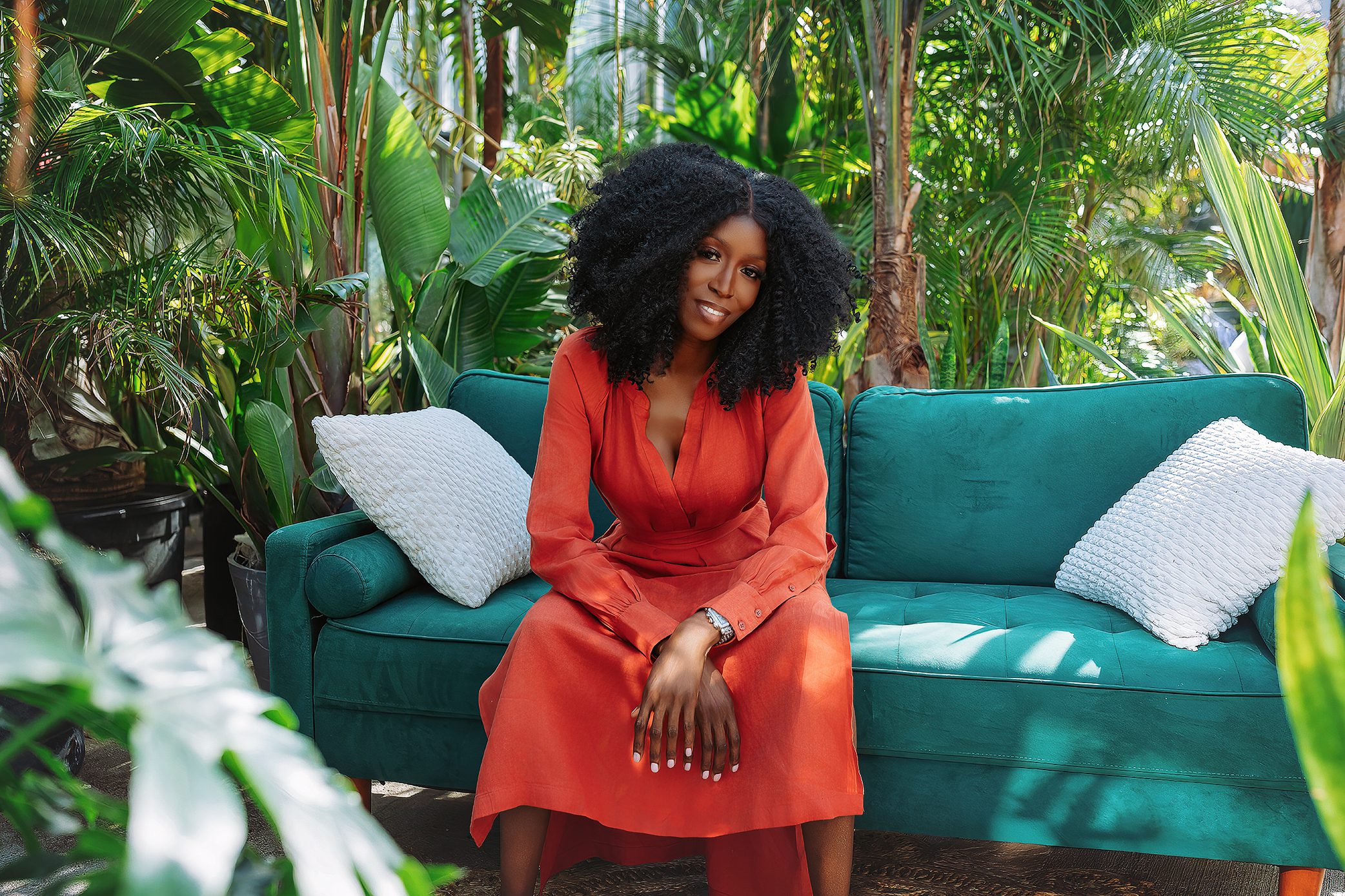 A woman with curly hair in an orange dress sits on a teal sofa with white pillows, surrounded by lush green plants in a bright, tropical setting.