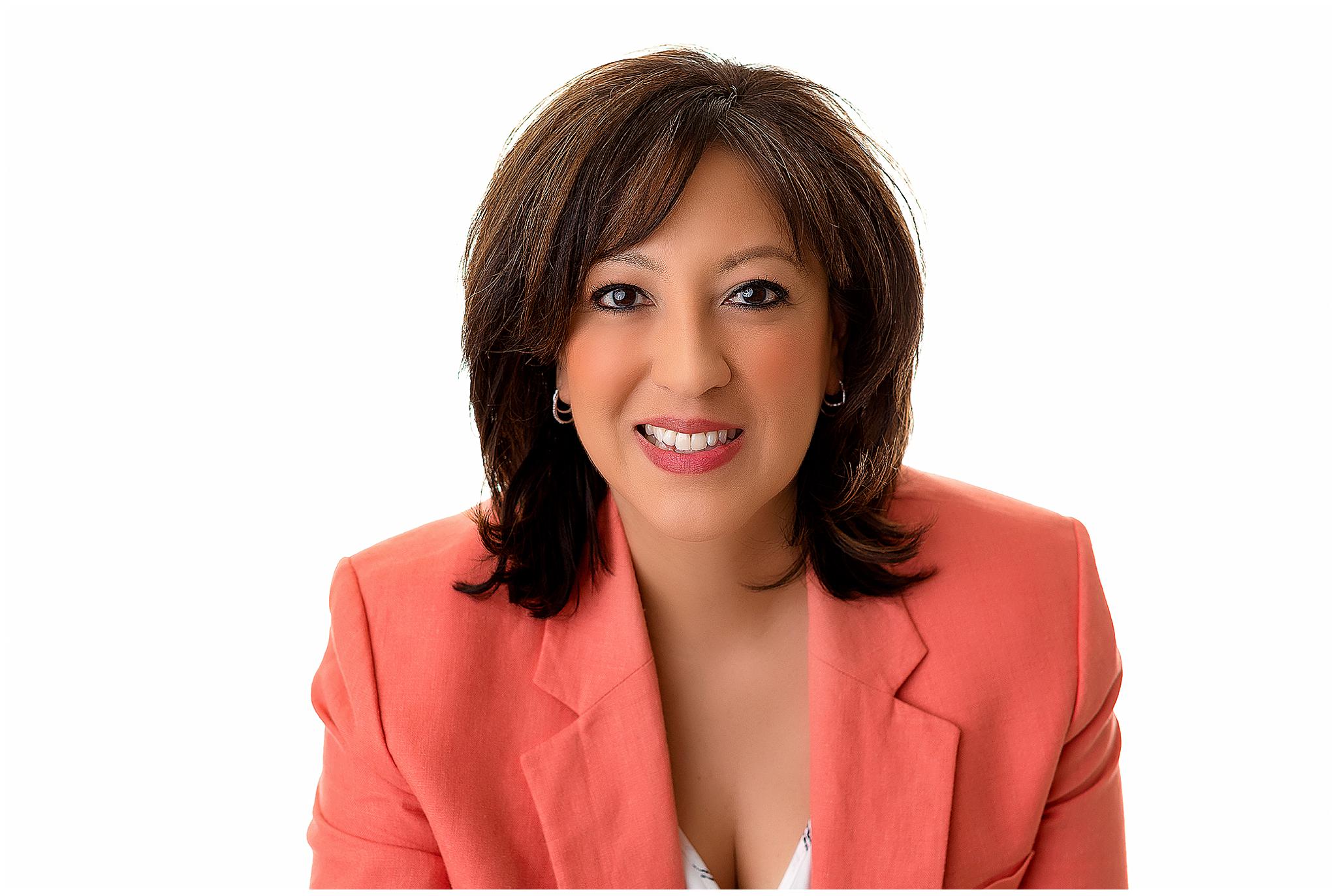 A woman with shoulder-length dark hair smiles in a headshot photography setting, wearing a coral blazer over a white top. She is pictured against a plain white background.